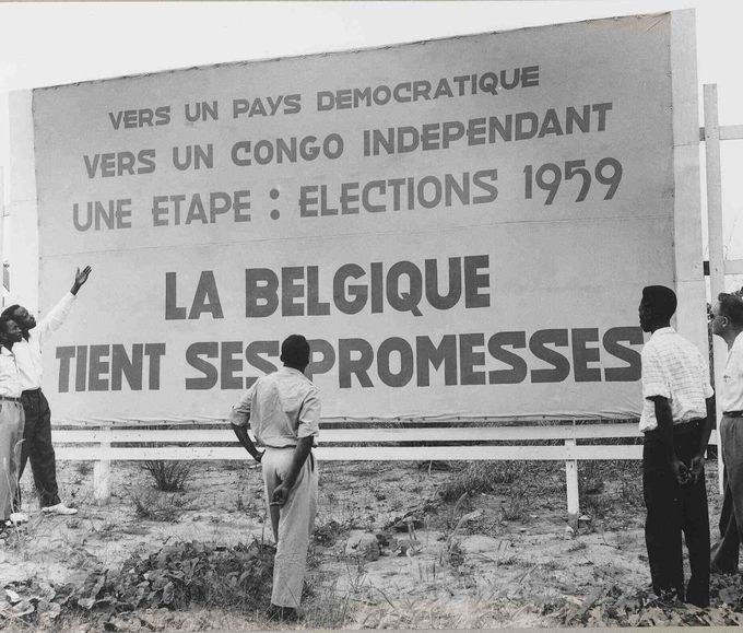 Photo of Congolese men and a colonial official in front of a poster for the provincial elections of December 1959 in the Belgian Congo