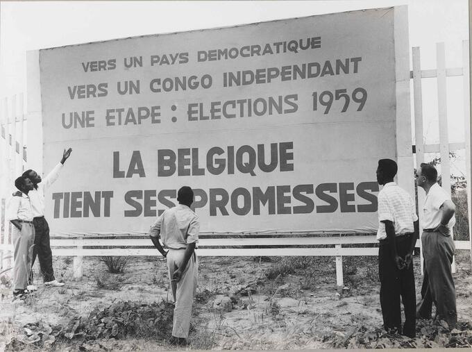 Photo of Congolese men and a colonial official in front of a poster for the provincial elections of December 1959 in the Belgian Congo